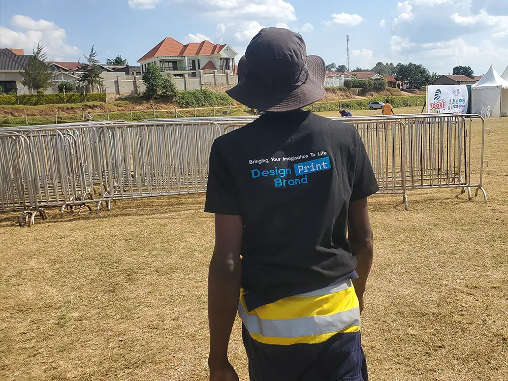 Team member near barrier setup — Kamog Artistry team member at the field barrier area during the Bushenyi setup process.